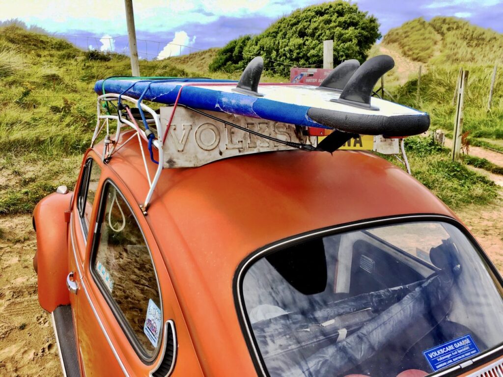Oranje VW kever met een surfplank op zijn dak bij de duinen