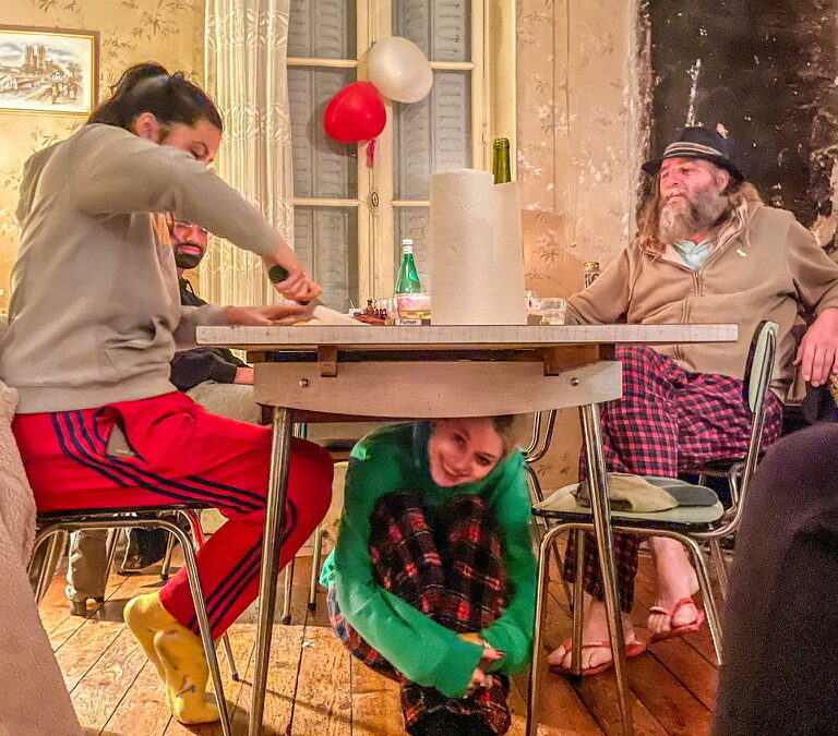 Meisje onder eettafel in groen T-shirt met twee mensen links en rechts aan de tafel zittend